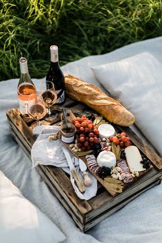 a wooden tray topped with bread and cheese next to two bottles of wine on top of a bed