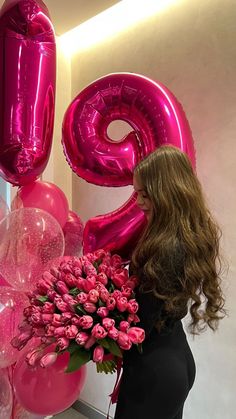 a woman standing next to a bunch of pink flowers and balloons in the shape of numbers