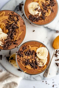 three desserts with bananas and chocolate on a white marble countertop next to spoons