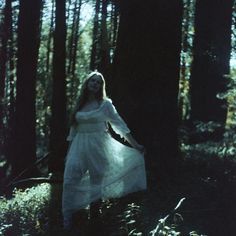 a woman in a white dress is walking through the woods with her arms spread out