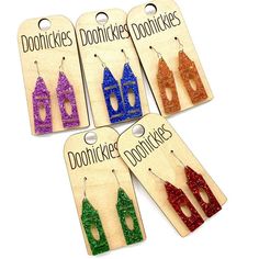 six pairs of earrings with different colors and shapes on wooden hangers, each featuring glittered houses