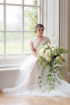 a woman sitting in front of a window holding a bouquet of white roses and greenery