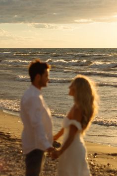 a man and woman standing on top of a beach next to the ocean at sunset