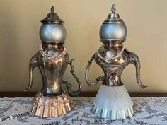 two silver vases sitting on top of a lace covered tablecloth with beads and pearls