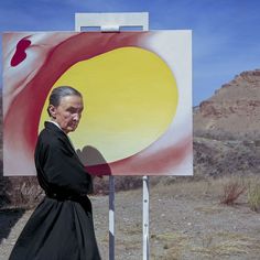 a man standing next to a large painting in the middle of a dirt field with mountains in the background