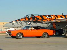 an orange car is parked in front of a fighter jet with the word canada painted on it