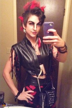 a woman taking a selfie with her cell phone in a bathroom mirror while wearing punk clothing