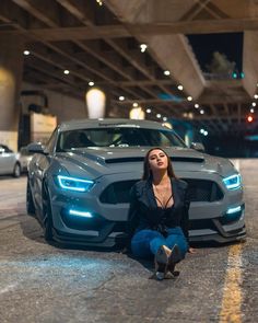 a woman is sitting on the ground next to a mustang car in a parking garage