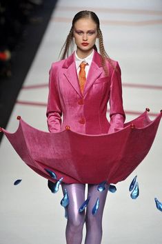 a woman in a pink suit and orange tie walks down the runway with an umbrella