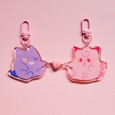 two pink and purple cat shaped keychains on a pink background, one is holding a heart