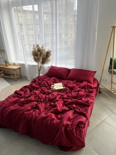 a bed with red sheets and pillows in front of a large window next to a plant