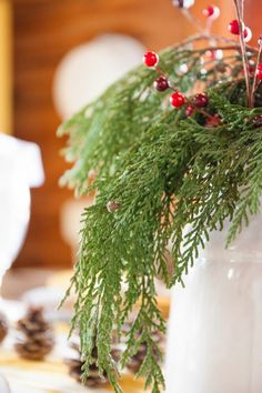 a white vase filled with greenery and red berries on top of a wooden table