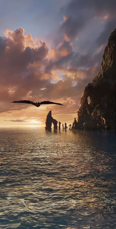 a large bird flying over the ocean near a rocky cliff and rock formation at sunset