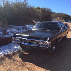 an old black car parked on the side of a dirt road in front of snow covered bushes