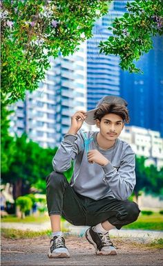 a young man squatting on the ground in front of a tree and tall buildings