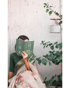 a woman sitting down reading a book in front of a plant with leaves on it