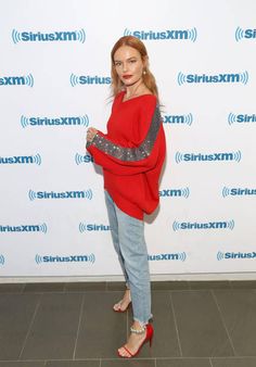 a woman in red sweater and jeans standing on tile floor next to wall with microphone