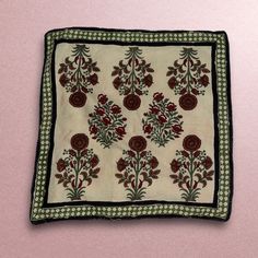 an embroidered square with red flowers and green leaves on it, sitting on a pink surface