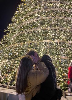 a man and woman kissing in front of a christmas tree with lights on it at night