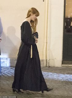 a woman in a long black dress is walking down the street while looking at her cell phone