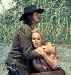 a man and woman sitting on top of a motorcycle in front of some tall grass