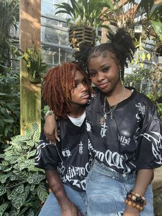 two young women sitting next to each other in front of plants and greenery, one is holding her arm around the other's shoulder