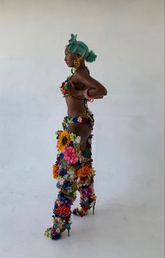 a woman with flowers on her body and headdress, standing in front of a white wall