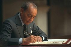 a man sitting at a desk writing on a piece of paper with a pen in his hand