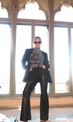 a woman standing in front of a window wearing black leather pants and a gray shirt