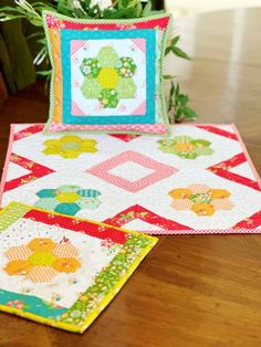 two quilts are sitting on a table next to a potted plant with flowers in the background