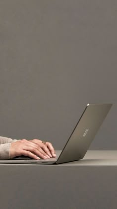 a woman is typing on her laptop while sitting at a table with a gray wall in the background