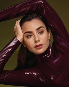 a woman with long dark brown hair wearing a shiny purple bodysuit and holding her hands on her head