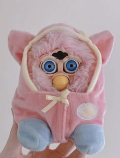 a hand holding a pink stuffed animal with big blue eyes and a hoodie on it's head