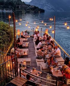 people are sitting at tables on the dock by the water with lights strung over them