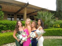 four beautiful young women standing next to each other in front of a house holding bouquets