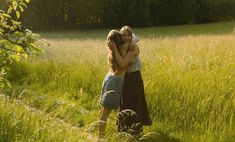 two women hugging each other in the middle of a field with tall grass and trees behind them