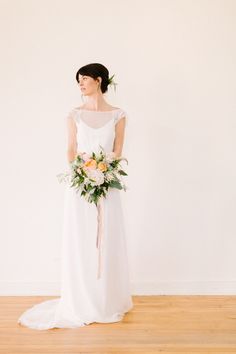 a woman in a white dress holding a bouquet and standing on a wooden floor with her back to the camera