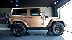 a tan jeep is parked in front of a large window with black rims on it