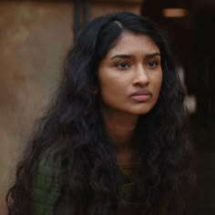 a woman with long black hair and green shirt looking off to the side while standing in front of a building