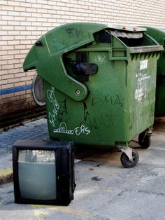 a tv sitting on top of a green trash can next to a black box with writing on it