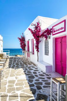 an empty street with tables and chairs next to the ocean in front of some white buildings