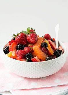 a white bowl filled with fruit on top of a pink table cloth next to a spoon