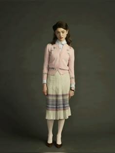 a woman standing in front of a gray background wearing a skirt and pink cardigan