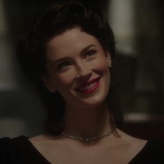 a smiling woman in a black dress with red lipstick on her face and necklace around her neck