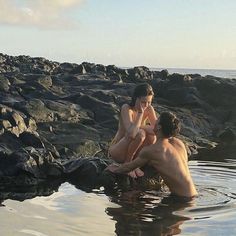 two people are sitting in the water near some rocks and one is holding a cell phone