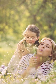 a woman and child laying in the grass with dandelions flying around their heads