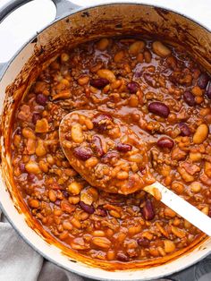 a large pot filled with beans and meat on top of a white cloth next to a wooden spoon