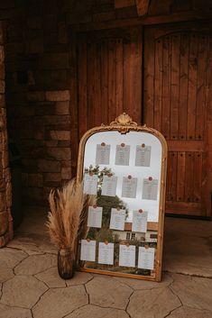 a wooden frame with seating cards on it next to a vase filled with dry grass