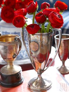 red roses are in a silver vase on a table with two cups and one is empty