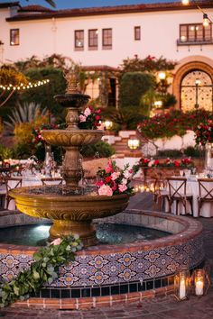 a fountain with flowers on it in front of a building at night, surrounded by tables and chairs